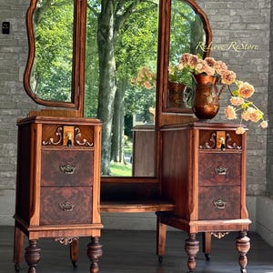 May include: A wooden vanity table with a tri-fold mirror, featuring two side cabinets with two drawers each, ornate metal pulls, and turned legs on casters. A copper-colored pitcher filled with artificial peach-colored flowers sits on top.