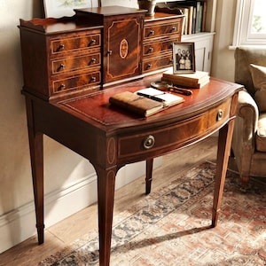 May include: A dark wood writing desk featuring a curved front drawer with a brass pull, a hutch with six small drawers, and a central cabinet door. The desktop surface is inlaid with a leather-like material and holds a leather-bound journal.
