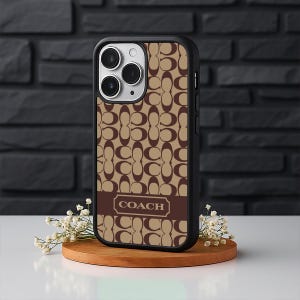 May include: A black iPhone case with a brown and tan repeating pattern. The case has a brown band across the bottom with the word "COACH" in a white rectangular frame. The phone case is on a wooden stand.