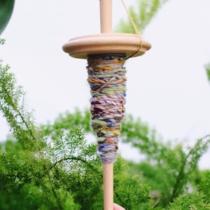 May include: A wooden spinning wheel with a colorful yarn skein wound around it. The yarn is a mix of purple, green, blue, and yellow.