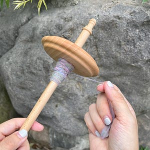May include: A wooden spinning wheel with a colorful yarn being spun. The yarn is a mix of pink, blue, and white.