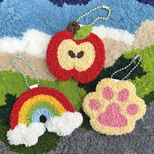 May include: Three colorful, textured keychains: a red apple with a tan center, a rainbow with white clouds, and a yellow paw print with pink pads. Each keychain has a beaded chain and a white tag.
