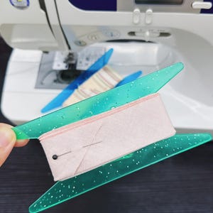 Pode incluir: Um projeto de costura em andamento, com uma fita de tecido rosa claro presa por uma guia verde transparente com manchas brancas. Uma máquina de costura está ao fundo, com uma guia azul e tecido listrado.