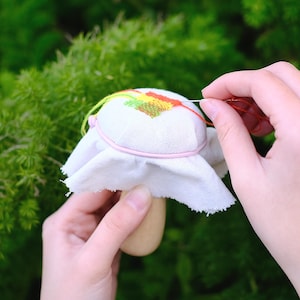Può includere: Un cuscino per spilli a forma di fungo in tessuto bianco con un motivo a punto croce arcobaleno. Il cuscino è cucito con un filo rosso.