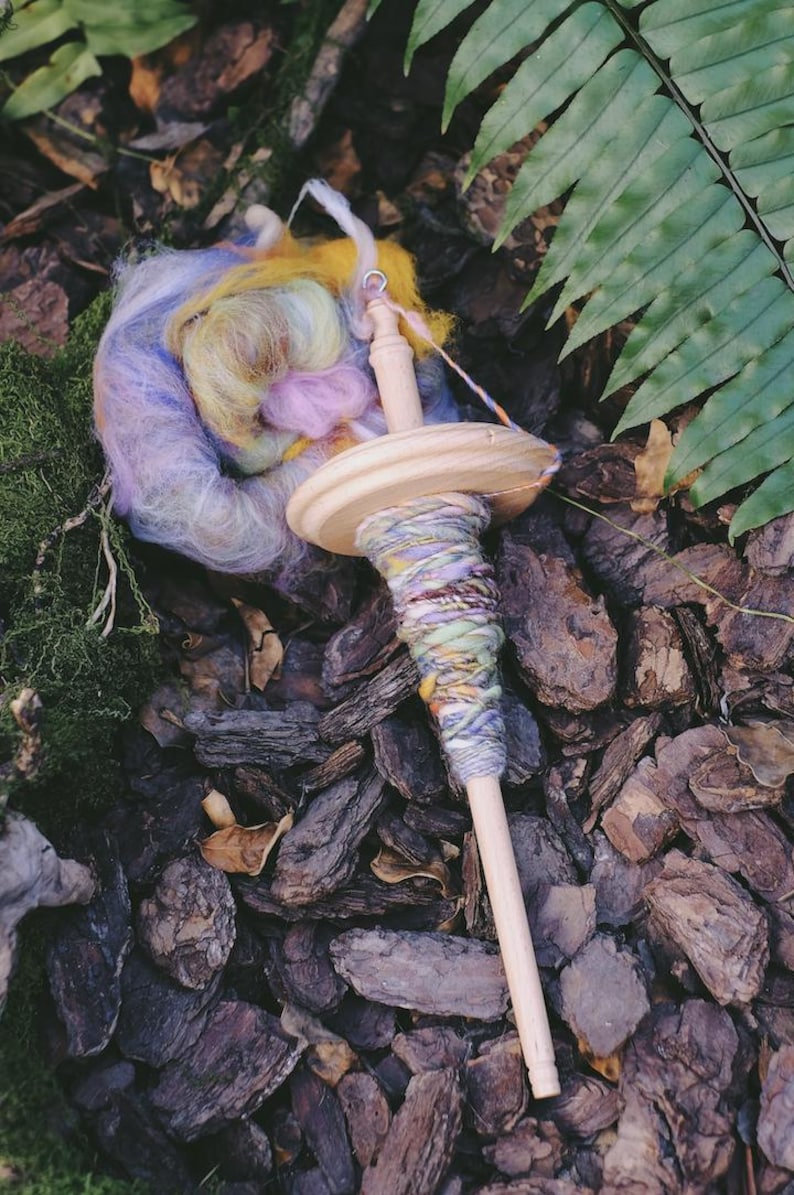 May include: A wooden spinning wheel with a colorful yarn ball and a skein of yarn. The yarn is a mix of purple, blue, yellow, and pink.