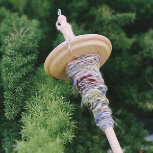 May include: A wooden spinning wheel with a colorful yarn skein wound around it. The yarn is a mix of purple, blue, green, and yellow.