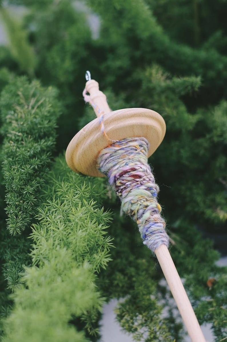 May include: A wooden spinning wheel with a colorful yarn skein wound around it. The yarn is a mix of purple, blue, green, and yellow.