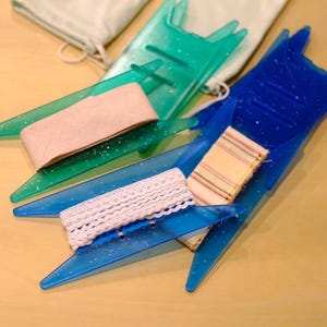 May include: Two translucent plastic yarn bobbins in teal and blue with glitter accents. The teal bobbin holds a solid light pink ribbon, while the blue bobbin holds a white lace trim and a striped fabric strip. A white fabric pouch is visible.