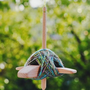 Op de afbeelding: Een houten drop-spindel met een bol veelkleurig garen. De spindel hangt tegen een wazige groene achtergrond. Het garen is een mix van blauw, groen en bruin. Een traditioneel hulpmiddel voor het handmatig spinnen van garen.