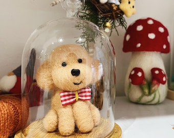 May include: A small, beige, felt poodle wearing a red and white bow tie is displayed under a glass dome on a wooden base.