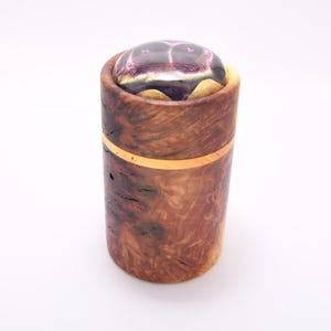 May include: A cylindrical wooden box with a decorative lid. The box is made of dark brown wood with a natural grain pattern and a gold-coloured band. The lid is a clear, rounded shape with purple and gold accents.