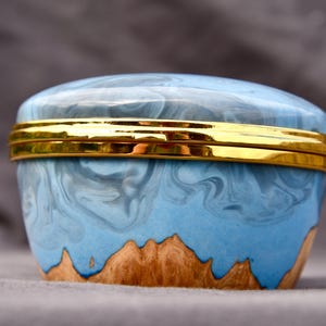 May include: A small round lidded container featuring a polished gold-tone metal rim. The body is crafted from a combination of natural light brown wood at the base and a smooth, marbled light blue resin top with swirling white patterns.