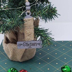 May include: A small artificial Christmas tree with a burlap base and a wooden ornament that says "Gibson" with paw prints. The tree is decorated with red and green jingle bells.