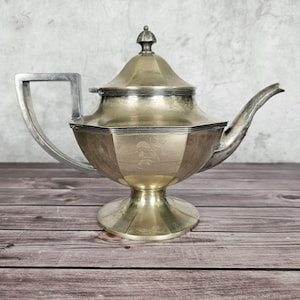 May include: A metallic teapot with a faceted, hexagonal body and a matching lid featuring a small finial. The teapot has a wide, angular handle on one side and a curved, tapered spout on the other, all supported by a flared, circular pedestal base.