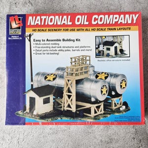 May include: A model train kit box featuring a National Oil Company scene. The box displays a miniature oil refinery with silver tanks, a wooden structure, and a small building. Text on the box includes "Easy to Assemble Building Kit."