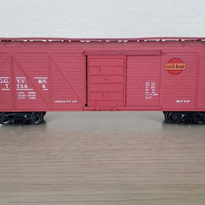 May include: A maroon model train boxcar with black wheels. The boxcar has the text "G.G. 7", "Y.V. 756", and "&N. 9" on the side. The words "Gulch Route" are also visible. The boxcar is sitting on a wooden surface.
