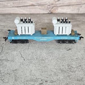 May include: A blue model flatcar train carriage labeled Great Northern 42764, featuring two light gray electrical transformer loads with black insulators and a small brown wooden crate secured on the flat deck, supported by black wheel bogies.