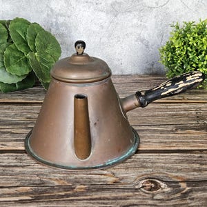 May include: A copper teapot with a conical body, a short spout, and a lid featuring a small black knob. The teapot has a side handle made of black wood with visible white paint wear and a weathered, oxidized patina on the metal surface.