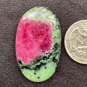 Pode incluir: Uma pedra preciosa de zoisite polida de formato oval com um centro rosa vibrante e bordas manchadas de verde e preto, posicionada ao lado de uma moeda de 25 centavos de dólar americano para mostrar o tamanho relativo da pedra.