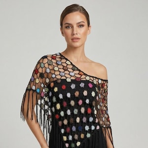 Op de afbeelding: Een zwarte gehaakte poncho met een zeshoekig netpatroon en veelkleurige, pluizige, ronde accenten in tinten rood, blauw, geel en roze. Het kledingstuk heeft een asymmetrische halslijn en lange zwarte franjes langs de onderkant.