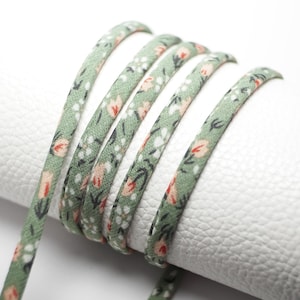 May include: Close-up of a spool of floral ribbon. The ribbon is a light green color with a repeating pattern of small pink and white flowers and dark green leaves. The ribbon is wrapped around a white surface.