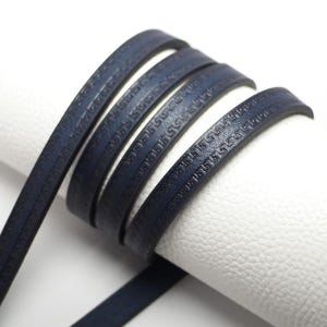 May include: Close-up of navy blue leather strips with a repeating geometric pattern along the edges. The strips are coiled on a white surface, showcasing the texture and detail of the material. The leather appears to be high-quality.