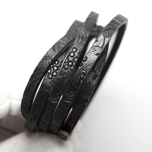 May include: A black leather wrap bracelet featuring a repeating embossed floral and circular pattern across the surface. The bracelet consists of multiple thin bands that overlap to create a layered appearance with a textured finish throughout.