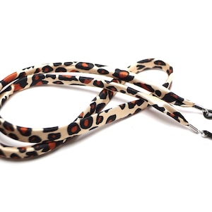 Può includere: Un cordino per occhiali con stampa leopardata con macchie nere e arancioni su sfondo crema. Le estremità hanno piccoli anelli di gomma nera per il fissaggio agli occhiali. Il cordino è ad anello e poggia su una superficie bianca.