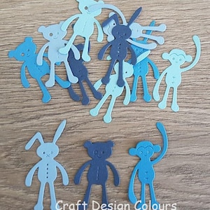 May include: A collection of die-cut paper shapes in various shades of blue, featuring silhouettes of teddy bears, rabbits, and monkeys. Each figure displays simple stitched-line details on the torso and face. The text Craft Design Colours is visible.