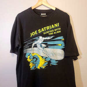 May include: Black t-shirt with a graphic of a silver surfer riding a surfboard with the text "Joe Satriani Surfing With The Alien" in yellow.