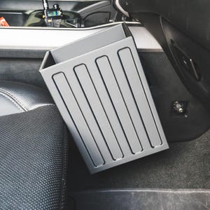 May include: A rectangular, light grey plastic storage bin with vertical ribbed detailing on the exterior sides. The container is positioned in the footwell area of a vehicle, resting between the centre console and the passenger side seat.