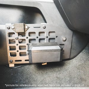May include: A matte gray metal MOLLE panel mounted to the side of a vehicle center console. The rectangular panel features a grid of laser-cut slots and circular holes, secured with black bolts. A small black rectangular box is attached to the panel.