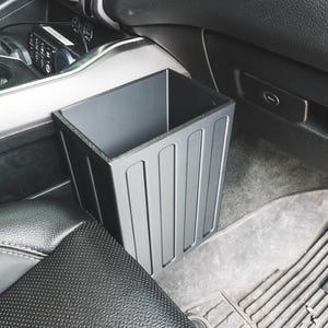 May include: A rectangular black car bin with vertical recessed panels on the exterior sides. The container is positioned in the passenger footwell area of a vehicle, featuring a hollow interior and a flat top rim for waste disposal.