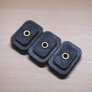 May include: Three black, rectangular objects with a textured surface and a gold-colored circular insert. The objects are arranged in a row, slightly overlapping, against a light brown background.