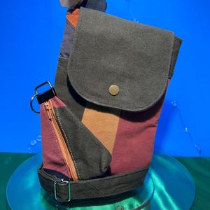 May include: A patchwork crossbody bag in shades of burgundy, brown, and grey. It has a flap closure, a small zipped pocket, and metal hardware. The bag features a strap and a metal clasp. The bag is designed to be worn across the body.