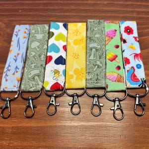 May include: Seven patterned fabric keychains with metal clasps. Designs include flowers, mushrooms, hearts, and ice cream. Each keychain is approximately 15 cm long, featuring a different colourful pattern.