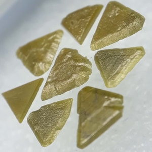 Può includere: Una collezione di otto piccole pietre preziose triangolari, traslucide e di colore verde chiaro. Le pietre preziose sono disposte in un motivo circolare su una superficie bianca.