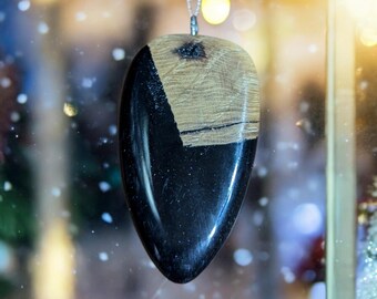 May include: A black and wood resin pendant with a teardrop shape hangs from a silver chain. The pendant has a unique design with a black resin base and a wood inlay.