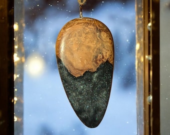 May include: A wooden and resin pendant with a brown wood top and a dark green and blue resin bottom. The pendant is shaped like a teardrop and has a silver chain.