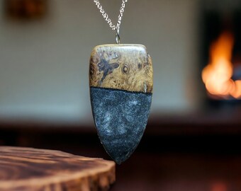 May include: A wooden and resin pendant with a silver chain. The pendant is shaped like a teardrop and has a dark gray resin bottom half and a light brown wood top half. The wood has a natural grain pattern.