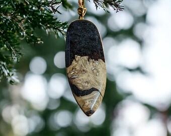 May include: A teardrop-shaped pendant made of wood and resin. The wood is a light brown with dark brown swirls, and the resin is a dark black. The pendant is hanging from a gold chain.