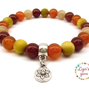May include: A bracelet made with orange, red, and yellow beads with a silver charm in the shape of a flower. The charm is attached to the bracelet with a silver chain.