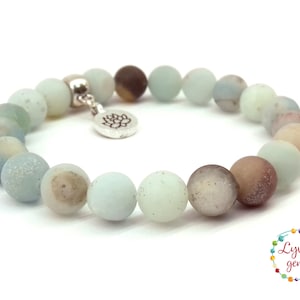 May include: A light blue and brown beaded bracelet with a silver charm featuring a lotus flower. The bracelet is made of natural stones and is approximately 7 inches in length.