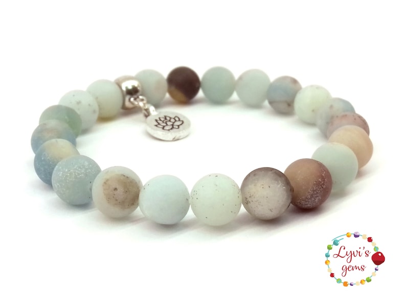 May include: A light blue and brown beaded bracelet with a silver charm featuring a lotus flower. The bracelet is made of natural stones and is approximately 7 inches in length.