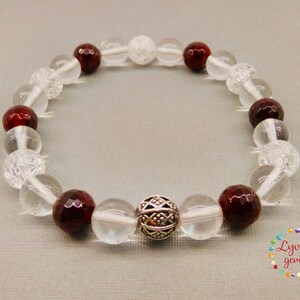 May include: A beaded bracelet featuring clear, faceted, and red-brown gemstones. The bracelet includes a decorative silver bead. The beads are strung on a clear elastic cord. The bracelet is displayed on a light gray surface.
