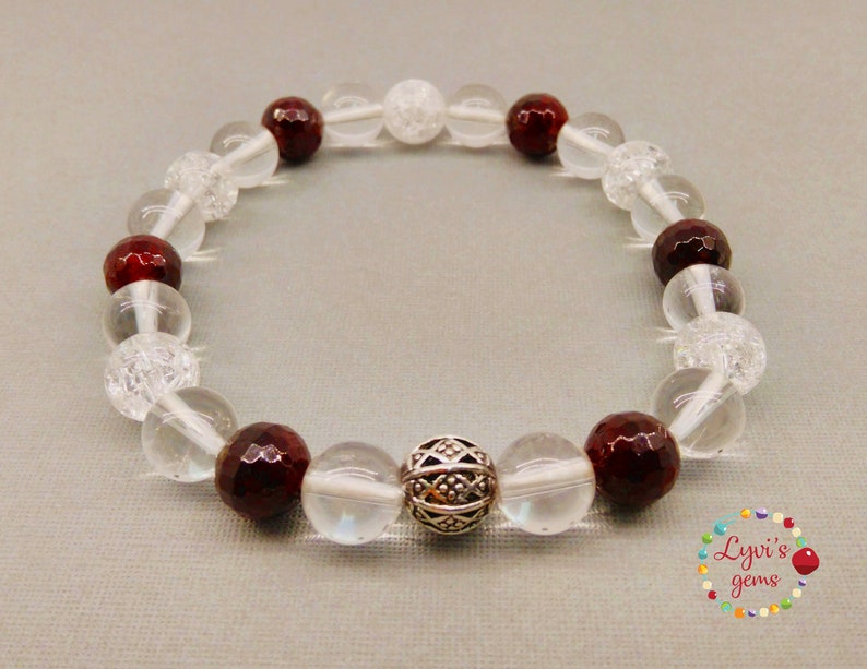 May include: A beaded bracelet featuring clear, faceted, and red-brown gemstones. The bracelet includes a decorative silver bead. The beads are strung on a clear elastic cord. The bracelet is displayed on a light gray surface.