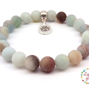 May include: A light blue, brown, and white gemstone bracelet with a silver lotus charm. The bracelet is made of round beads and is approximately 7 inches in length.