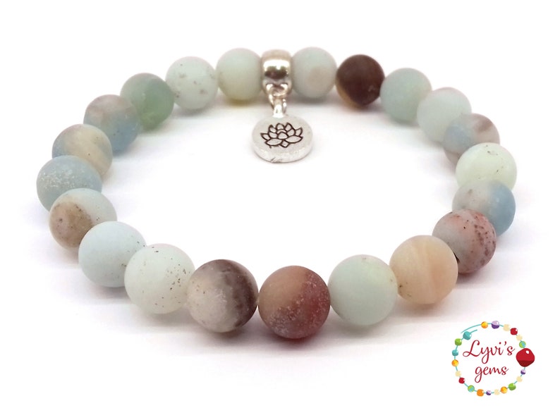 May include: A light blue, brown, and white gemstone bracelet with a silver lotus charm. The bracelet is made of round beads and is approximately 7 inches in length.