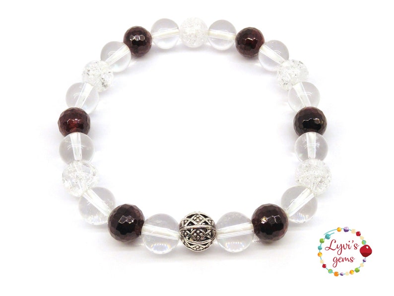 May include: A beaded bracelet featuring clear, faceted, and dark red gemstones. The bracelet includes clear, round beads and a decorative silver bead. The bracelet is on a white background. The image also includes a logo with the text "Lyvi's gems".