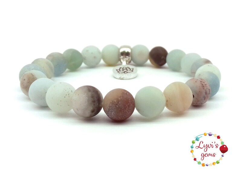 May include: A light blue, brown, and white gemstone bracelet with a silver lotus charm. The bracelet is made of round beads and is approximately 7 inches in length.